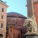 Statue of a carved white elephant and obelisk with a brown curved building behind and two coloured buildings either side