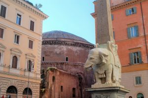 Statue of a carved white elephant and obelisk with a brown curved building behind and two coloured buildings either side