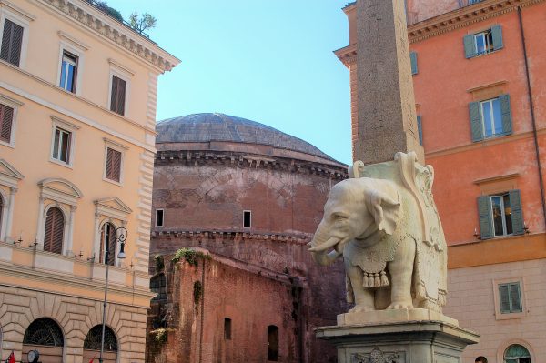 Statue of a carved white elephant and obelisk with a brown curved building behind and two coloured buildings either side