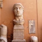 An ocre coloured wall with parts of an ancient Roman statue in front - head on a plinth, arm, knee and hand with finger pointing upwards