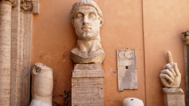 An ocre coloured wall with parts of an ancient Roman statue in front - head on a plinth, arm, knee and hand with finger pointing upwards