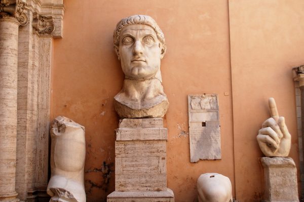 An ocre coloured wall with parts of an ancient Roman statue in front - head on a plinth, arm, knee and hand with finger pointing upwards