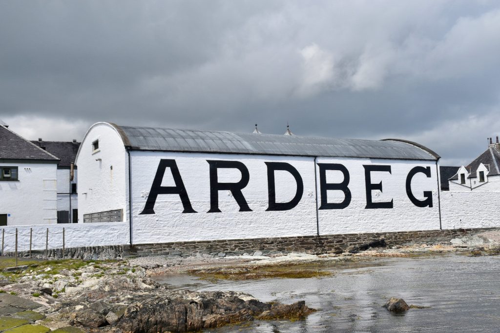 A stormy grey sky in the top of the photo. A body of still water surrounded by rocks at the bottom, In the middle is a white washed building with the word Ardbeg in large black letters on the front.
