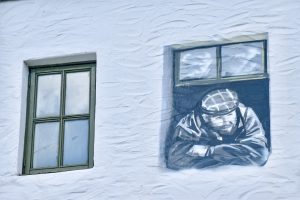 White washed wall with a green framed window to the left and a mural of a man looking out of a window to the right. The man in the mural is waring a hacker and flat cap, with a beard. HIs arms are crossed and he is looking down to his left.