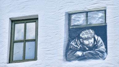 White washed wall with a green framed window to the left and a mural of a man looking out of a window to the right. The man in the mural is waring a hacker and flat cap, with a beard. HIs arms are crossed and he is looking down to his left.