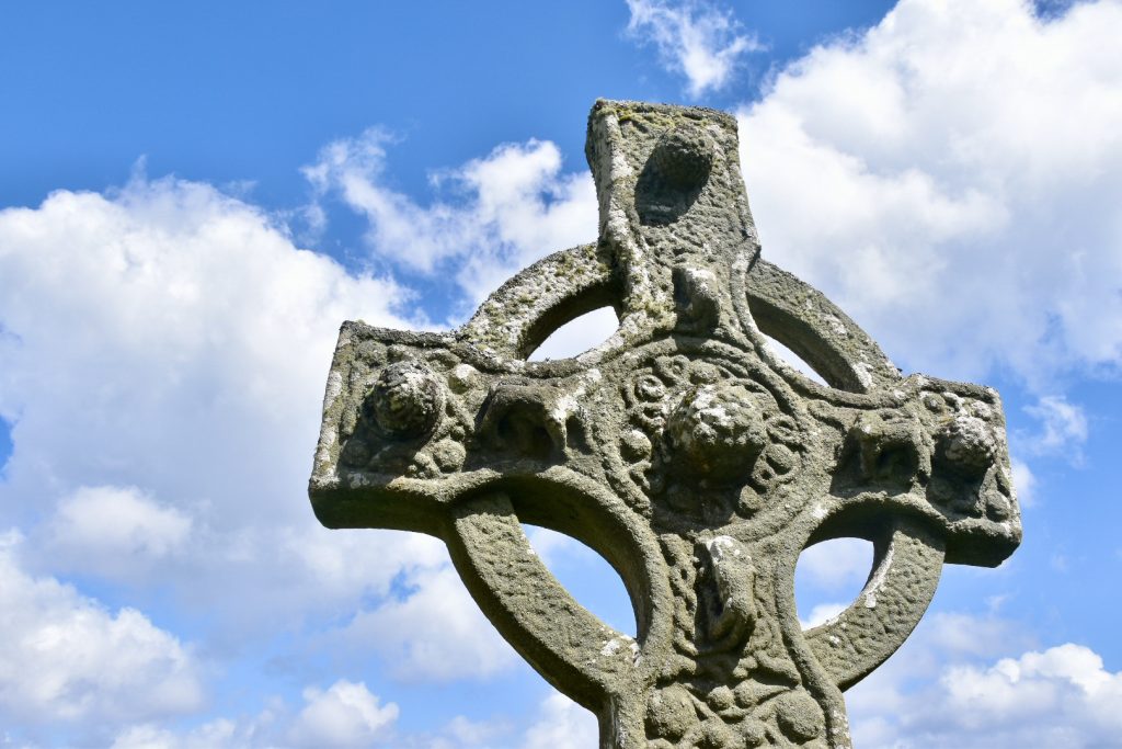 A blue sky in the background with fluffy white clouds. A ringed stone cross rises up from the bottom right of the frame. It has Celtic designs on it