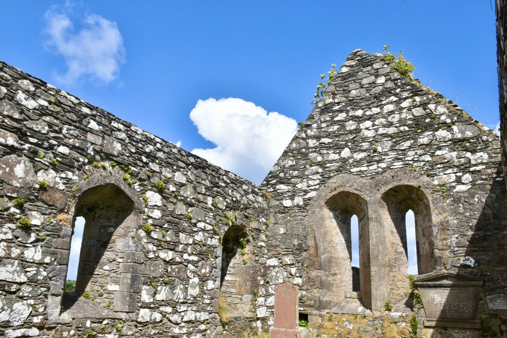 view upwards towards the end of an old stone building which looks like a church from the tombstone inside and arched windows. There is no roof, just a blue sky above and one patch of cloud.
