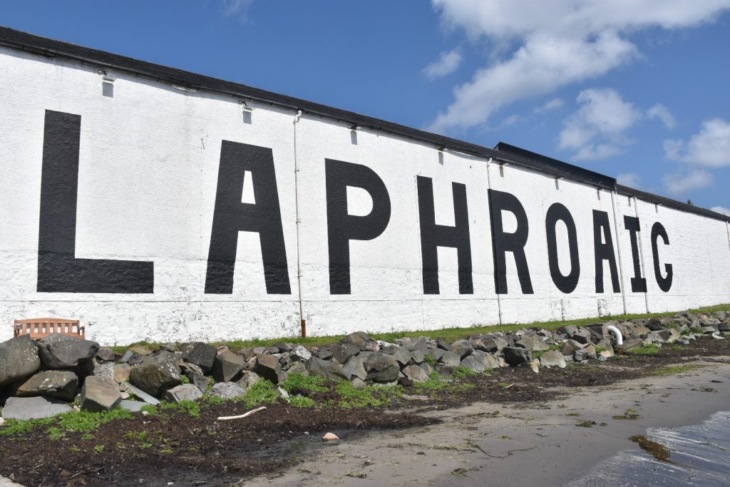 Blue sky at the top with some patchy white cloud. Rocky shore land with wet sand and a hint of water at the bottom. Across the middle is a white washed building with the words LAPHROAIG across the middle in black