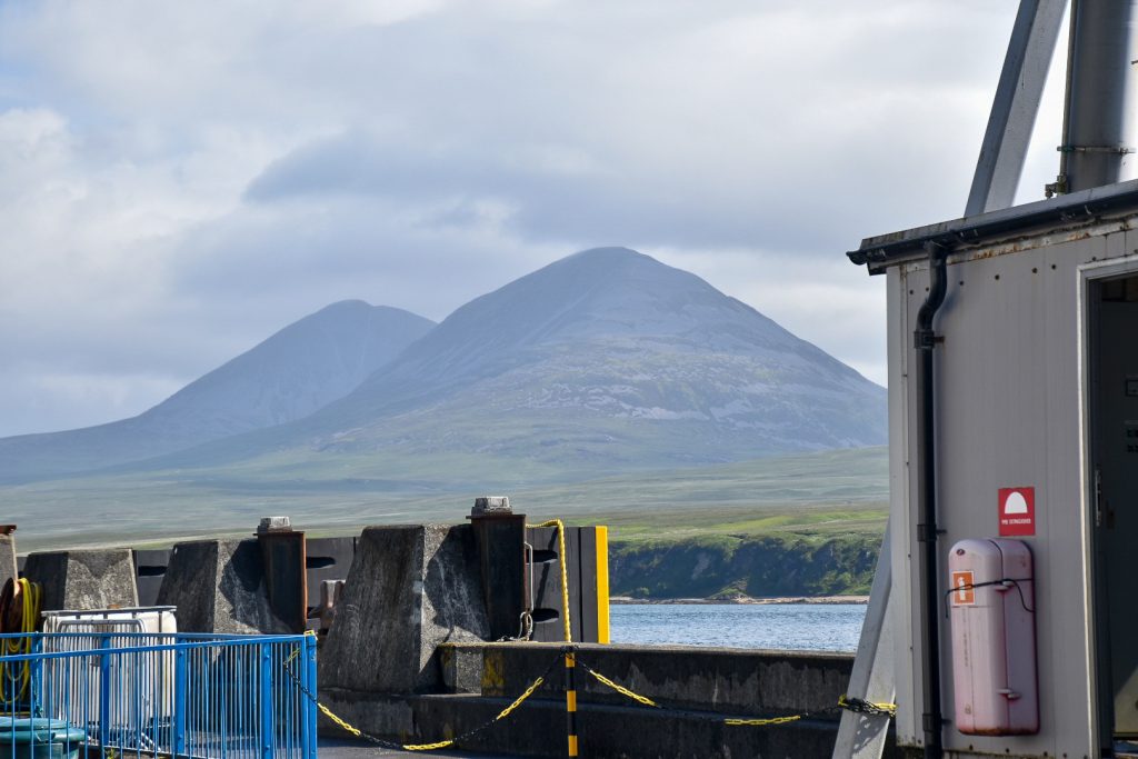 View of two peaked green mountains under a pale grey cloudy sky. Shot taken from what looks like a ferry on the water. Sides of the ferry in foreground