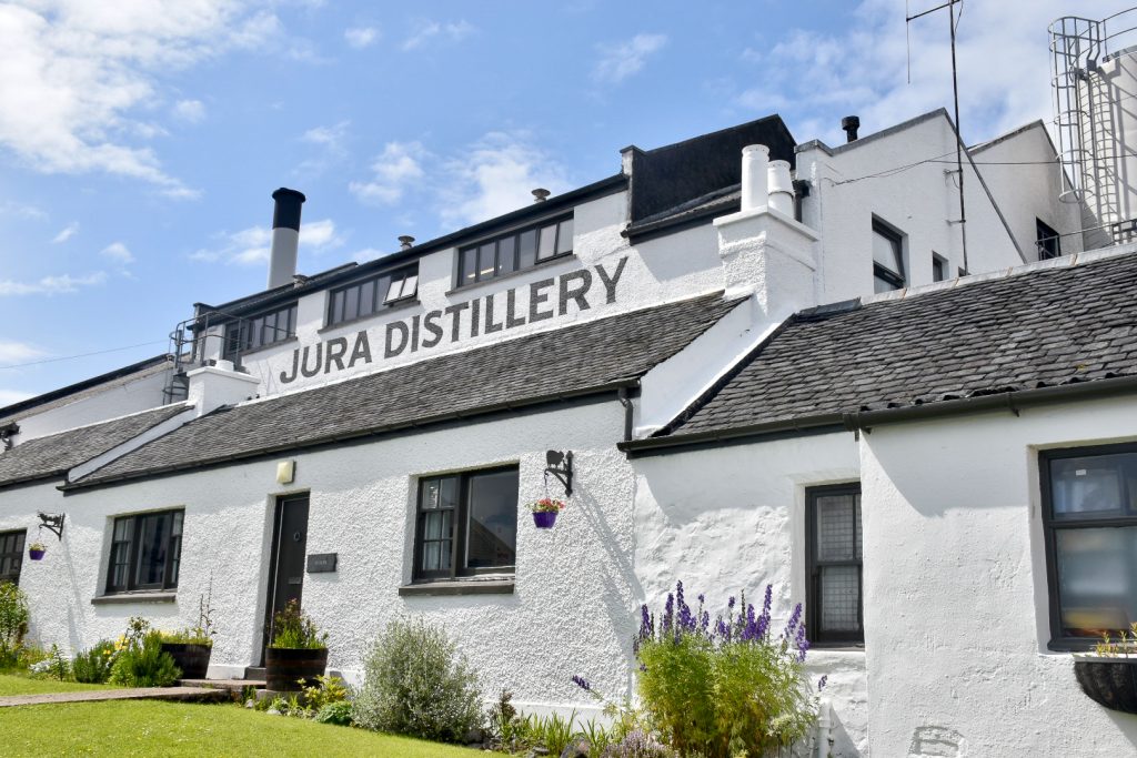 White building crosses the whole photo. Building has black slate roofs and the words Jura Distillery painted on the side. Green grass in front and blue sky with white clouds behind
