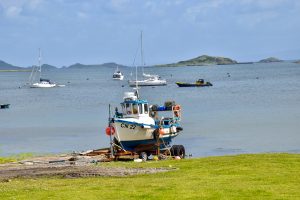 Landscape scene of a small white fishing boat on the grass with the water behind it and sailing boats on the water. Hills on the horizon. The light is dark grey