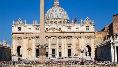Large impressive white stone building behind a square filled with people. Basilica behind the building and obelisk in front