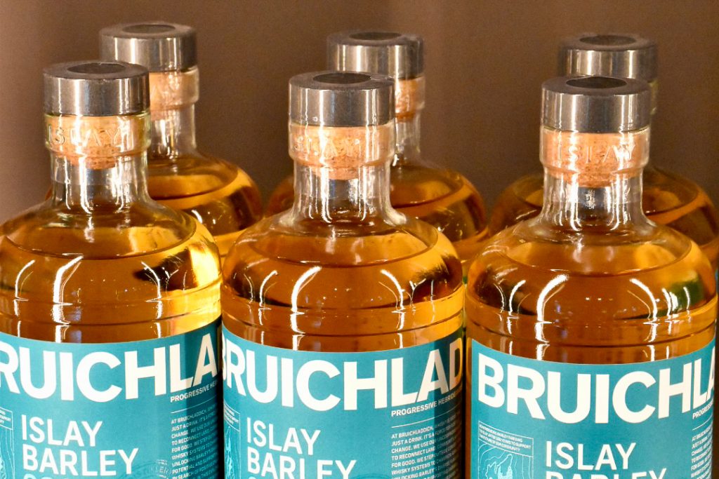 Row of 3 bottles of whisky across the middle of the frame, and a second row of 3 bottles behind. Each bottle has a turquoise label with white writing - Islay Barley, Bruichladdich