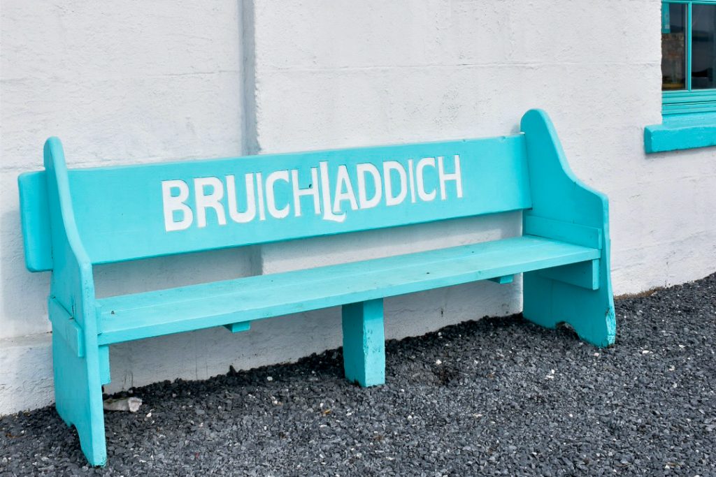A turquoise wooden bench sits on grey gravel in front of a painted white wall. The bench says Bruichladdich on the back support. Hint of a turquoise window frame in top right of frame