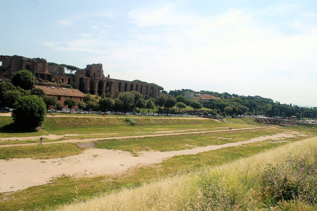 Looking along a long expanse of grass with trees on the horizon and old buildings. Pale sky above