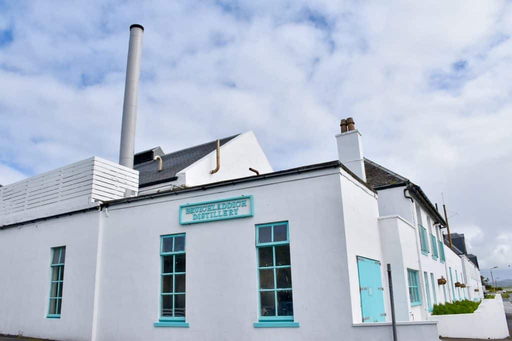 White washed building stretching across the bottom of the frame. Cloudy sky with a hunt of blue in the background. The building as turquoise framed windows, doors and a sign saying Bruichladdich Distillery. It has a thin silver chimney