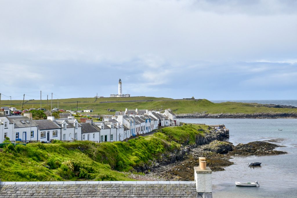 Top half of photo shows a grey sky with white cloud. The bottom of the photo features a row of small which cottages positioned around a small harbour. A white lighthouse stands on the land on the horizon. A couple of small boats float in the water.