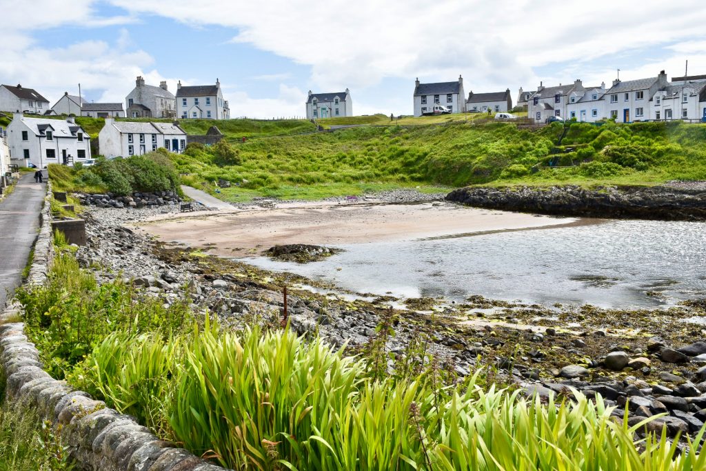 A row of white cottages surrounds a small harbour with rocky shore and walls around the edge. The houses are perched on a grassy bank. White cloud above with a small patch of pale blue sky
