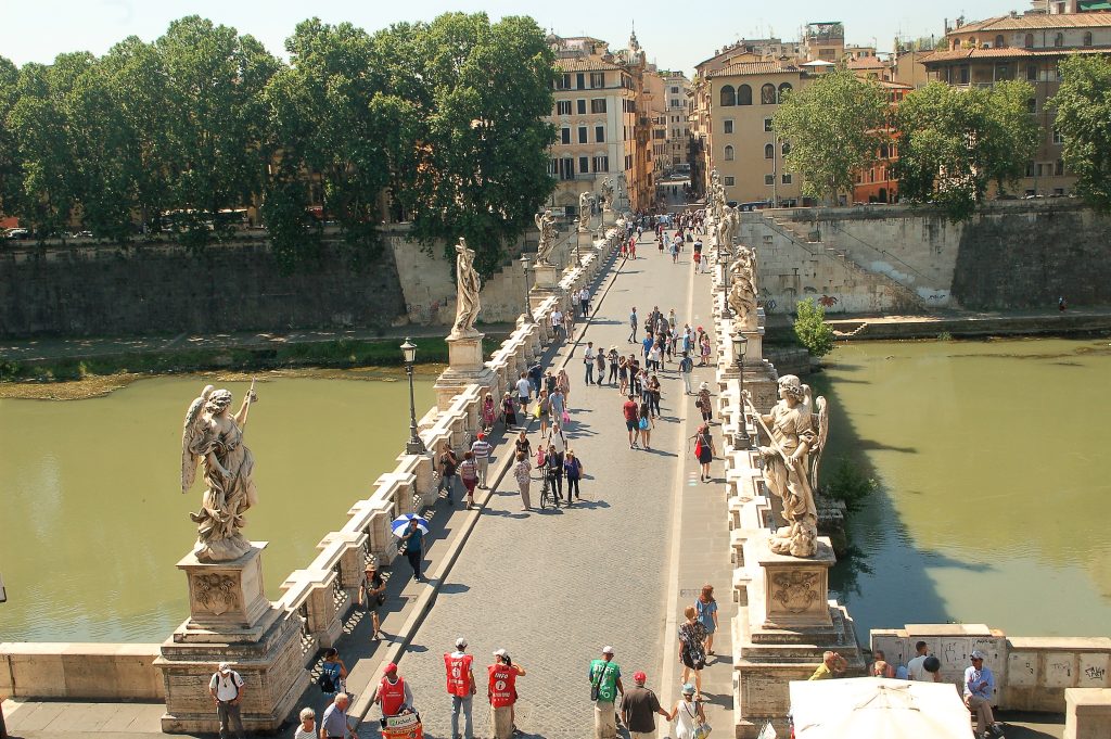 View looking down on a white bridge flaked with white marble statues over a green river. Trees and buildings on the other side of the river