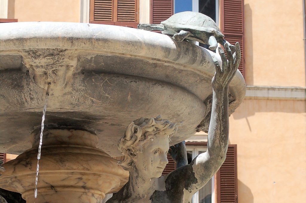 A stone water fountain fils the frame to the left hand side. It is supported by a stone figurine under the water bowl, reaching their arm to the top where a stone turtle is balanced on the rim. A small spout of water comes from a carved face to the left of the structure. Salmon pink building behind with brown shutters