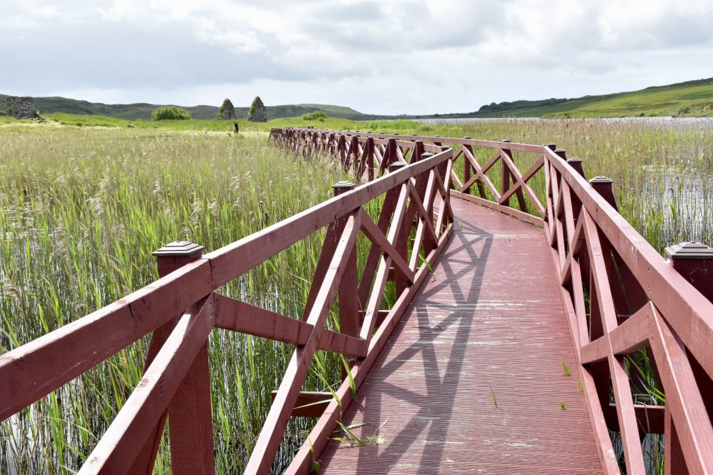 A reddish-brown wooden boardwalk with sides leads from the bottom of the photo towards the middle of the photo over a bed of reeds on water. Green countryside on the horizon with a pale grey sky above