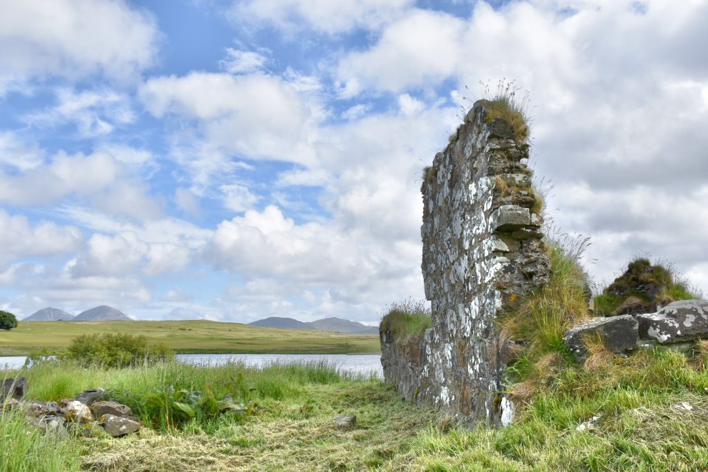 Top two-thirds is a blue sky filled with fluffy white clouds. There is a strip of green grass across the bottom of the frame and the ruins of an old stone wall to the right hand side. On the horizon you can see a number of hills across a body of water