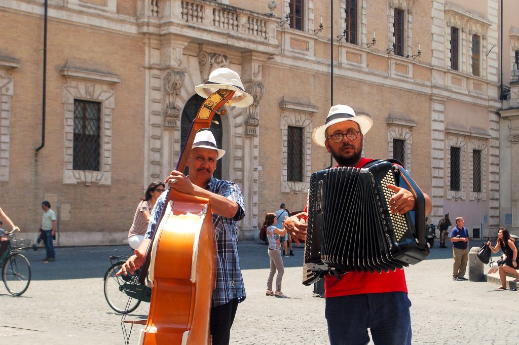 Two buskers face the camera with beautiful old white buildings in the background. The man on the left is playing the double bass with a straw hat on its arm, and the man on the right is playing the accordion. Both wearing straw hats