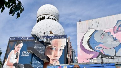 Cloudy blue sky in background. White golf ball structures in front with buildings at the bottom decorated with graffiti