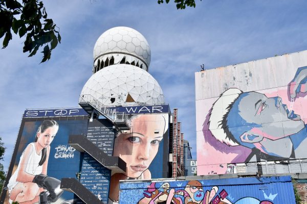 Cloudy blue sky in background. White golf ball structures in front with buildings at the bottom decorated with graffiti