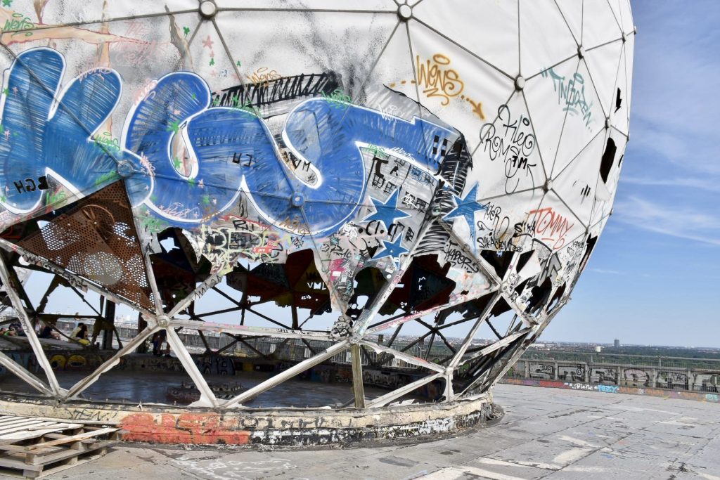 A large golf ball structure made of metal frame and covered in canvas sits on a concrete rooftop overlooking a forest and cityscape. The canvas on the frame is frayed and missing at the bottom, with graffiti on the lower parts.