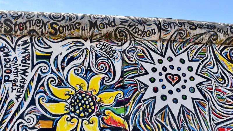 View of a painted wall with blue sky above. The wall is white with black flower outlines filled in with colours