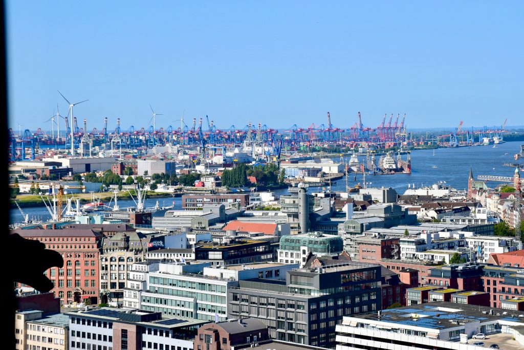 View from high across roof tops towards a dock full of red cranes and windmills (wind farm style). Water in the middle of the buildings. Blue sky above