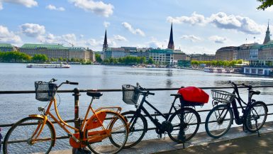 Row of three bikes leaning against black railings with area of water behind. On the other side of the water is a line of smart buildings and three church spires. Pale blue sky above
