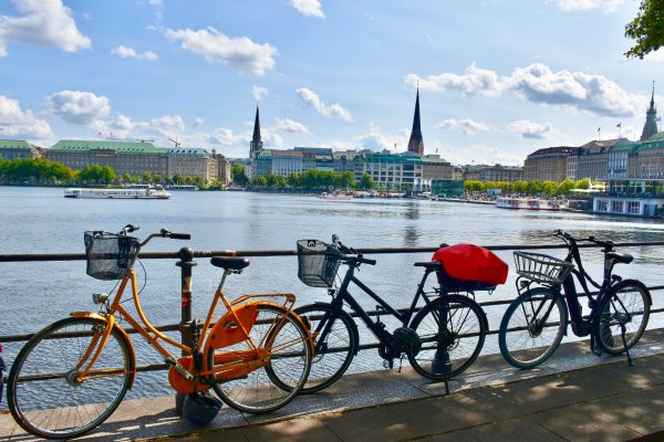 Row of three bikes leaning against black railings with area of water behind. On the other side of the water is a line of smart buildings and three church spires. Pale blue sky above