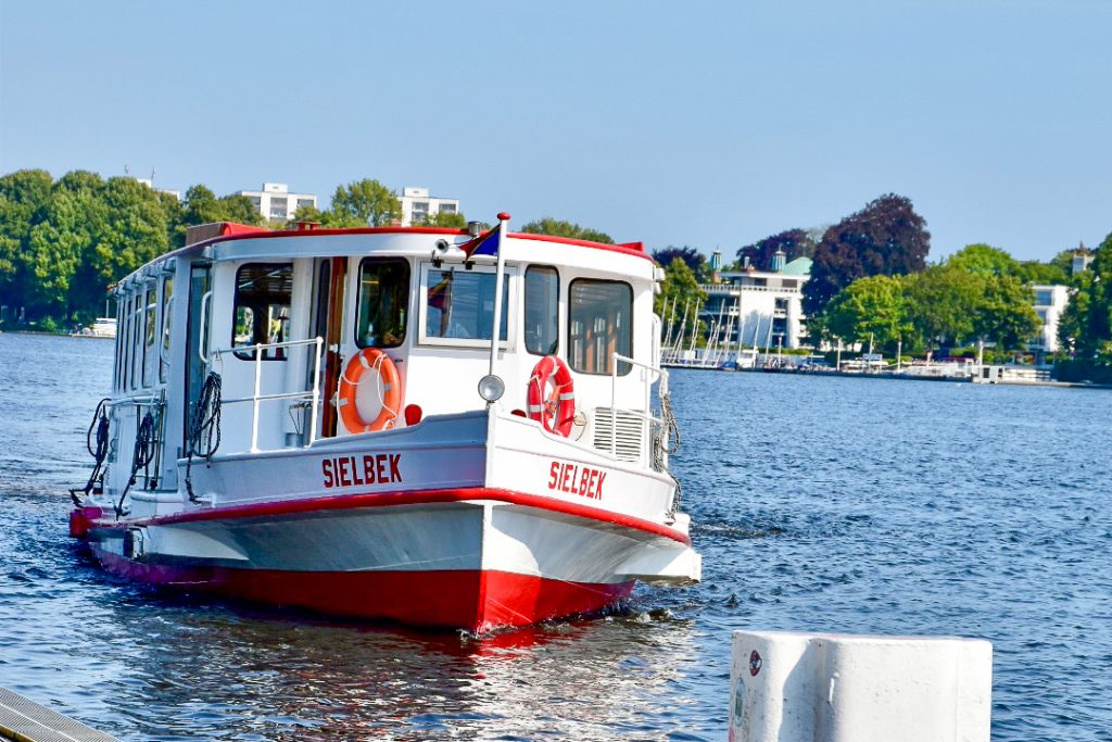 White barge style boat with red trim heading towards the camera. It is called Sielbek. Trees, buildings and sail boats in the distance behind. Pale blue sky