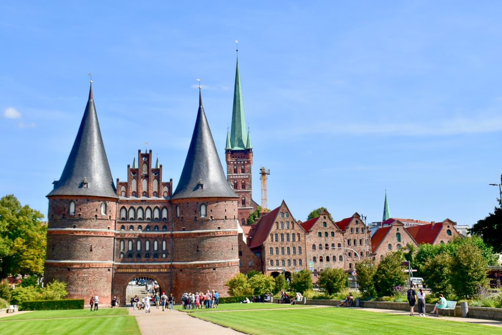Twin red brick circular towers with grey spires, linked together by a stepped gable building. Row of warehouse buildings to the right with stepped gable roofs. Lawn in front. Turquoise spire behind. Blue sky