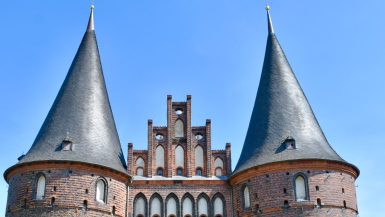 Close up of two circular red brick towers joined by stepped gable brickwork in between. Two circular spires in grey tiles above. Blue sky behind