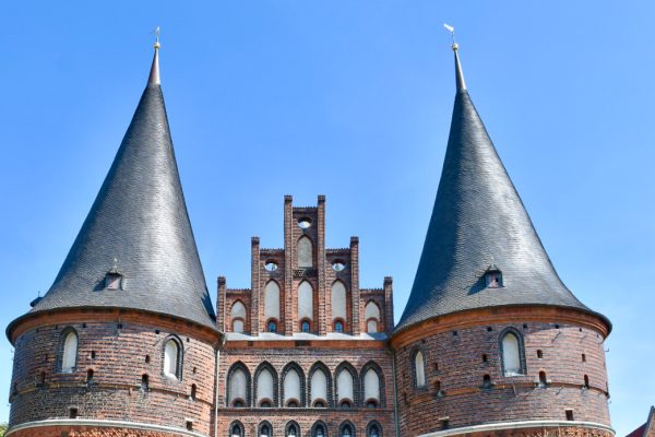 Close up of two circular red brick towers joined by stepped gable brickwork in between. Two circular spires in grey tiles above. Blue sky behind