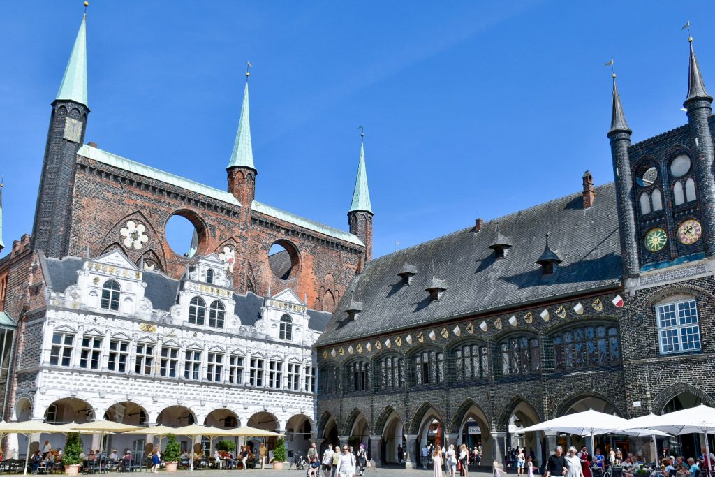 Pale blue sky above Medieval buildings on the corner of a square filled with tables and parasols. Ornate white building to the left with tall red brick structure behind with 3 turquoise towers on it. A low rise black stone building with shields to the right