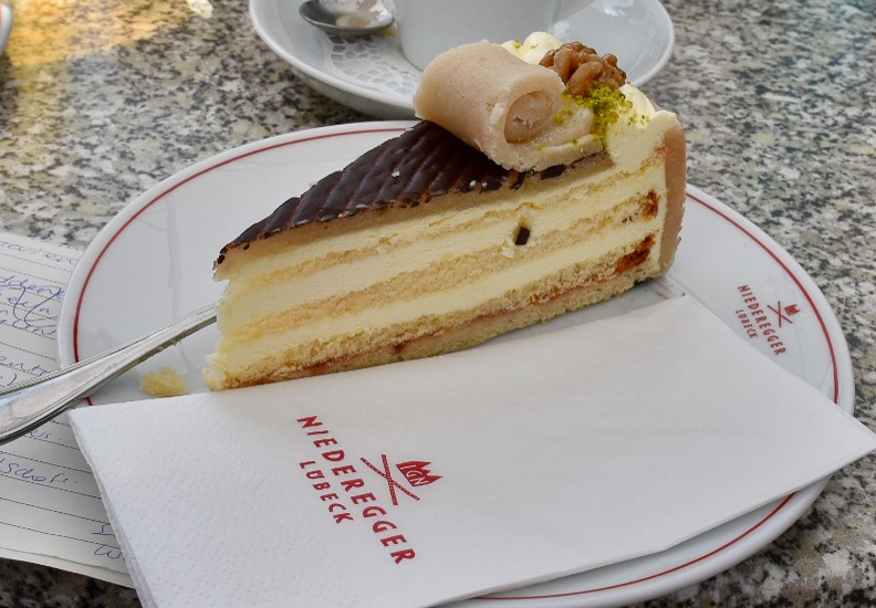 A slice of layered gateau filled with cream, sponge and chocolate sits on a white plate with red trim. It says Cafe Niederegger on it and the white napkin to its right. Cup of coffee behind