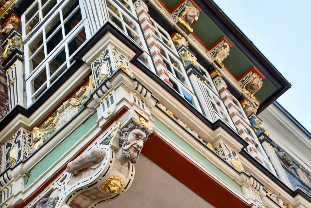Looking upwards at a very colourful carved building with a window sticking out from the main walls. Held up by painted male figurehead. Gold, green, orange, black features