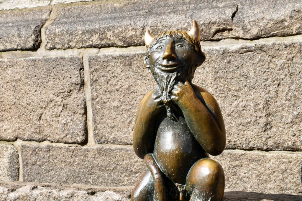 A small brass sculpture of a smiling devil with horns and tail sitting in front of a brick wall