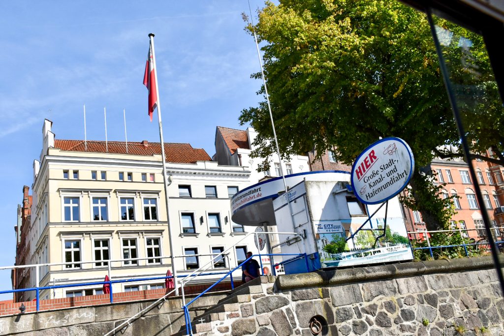 View of a stone quayside with a white boat ticket office at the top of a flight of stairs. Row of smarter buildings behind and trees to the right