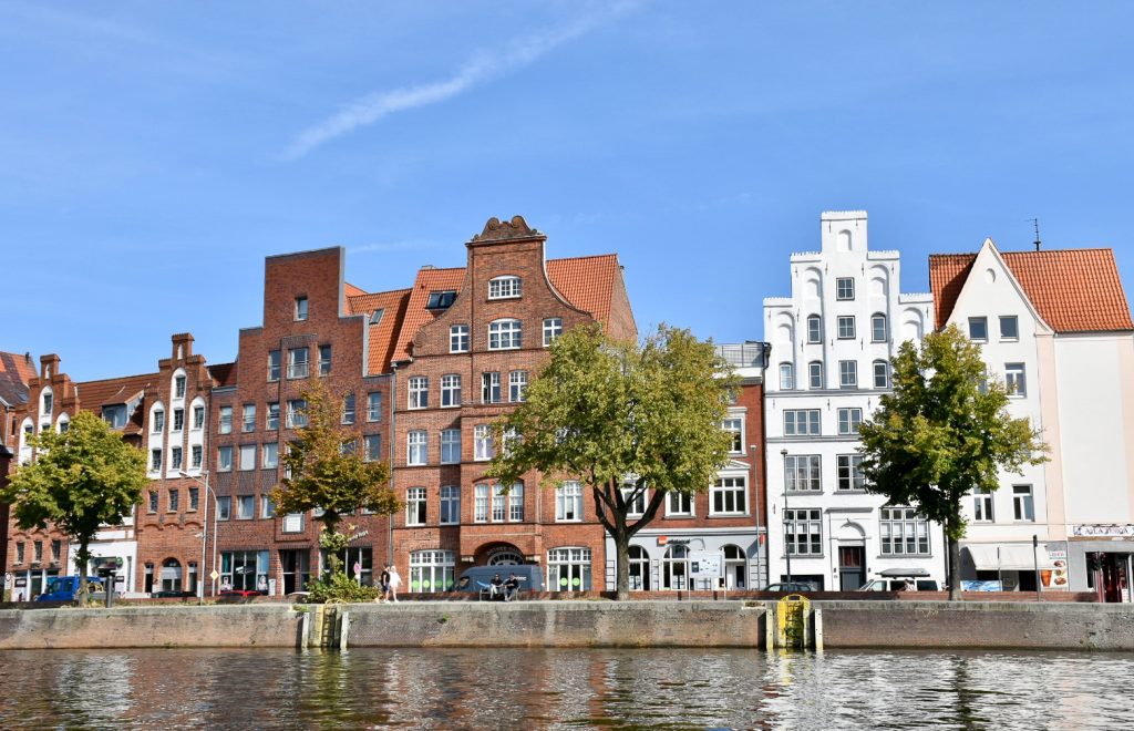 Row of old brick buildings with stepped gable roofs along a canal side. The buildings to the left are red and the two to the right are white. Blue sky above