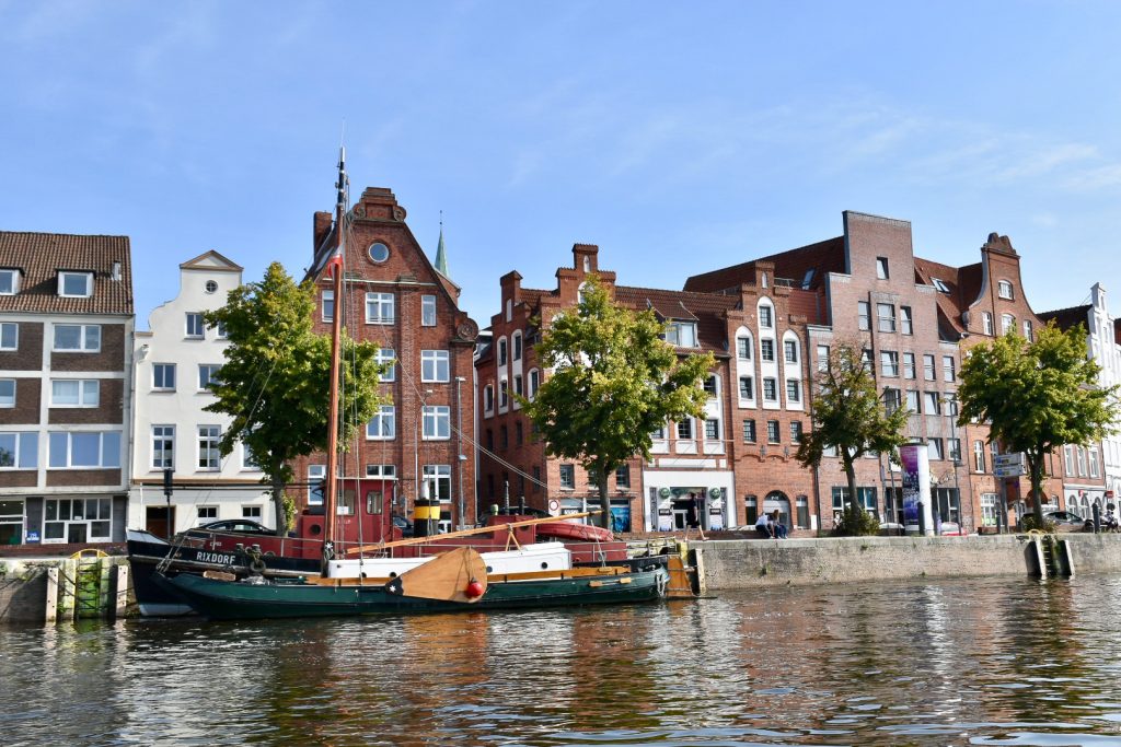 Row of old brick buildings with stepped gable roofs along a canal side. A green wooden sailboat is moored in front.