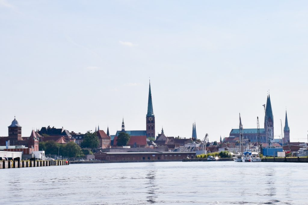 View along a body of water like a wide Canal or rover to Medieval town buildings at the end. There are several spires silhouetted against a pale sky