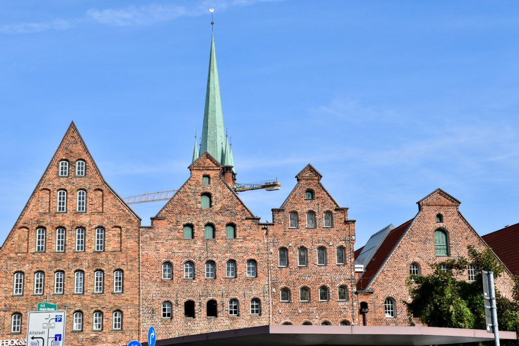 Row of 4 red brick warehouses with stepped gable roofs across the photo. The warehouses have 5 storeys each filled with arched window. Turquoise spire behind. Blue sky above
