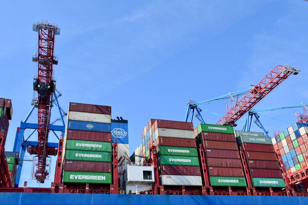 Rows of containers rise up from a ship. Crane to the left and right. Blue sky above.