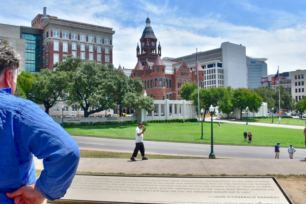 Man in blue shirt stands in bottle left of frame with hands on hips reading a sign about John Fitzgerald Kennedy. It overlooks a road and grassy area with American buildings beyond