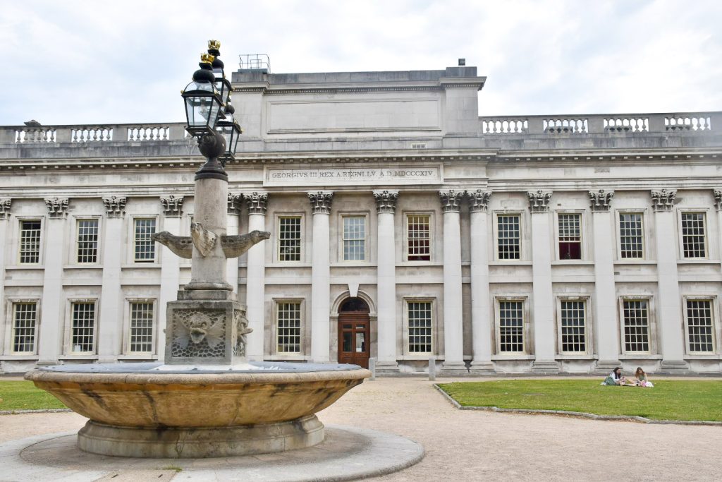 long white stone building with columns which reaches from one side of the photo to the other. It has rows of square pane windows over two flours. Grey sky above. Lawn in front with a stone water fountain to the bottom left